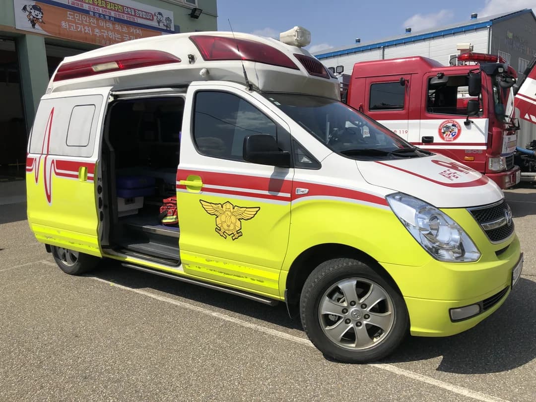 A 119 ambulance with its side door open, with a red fire engine behind it